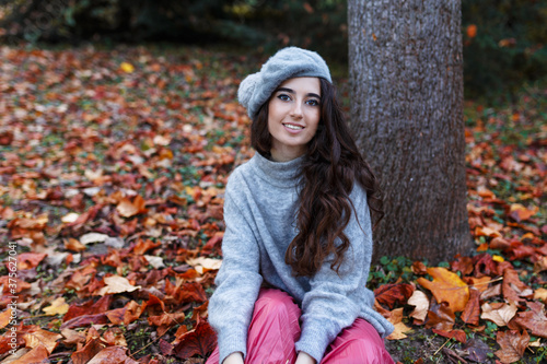 The young woman dressed in a beret and autumn clothes is sitting on the leaves near to a tree in the autumn season.