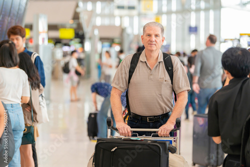 Handsome senior man with backpack walking with a luggage at airport terminal and airport terminal blurred crowd of travelling people on the background.