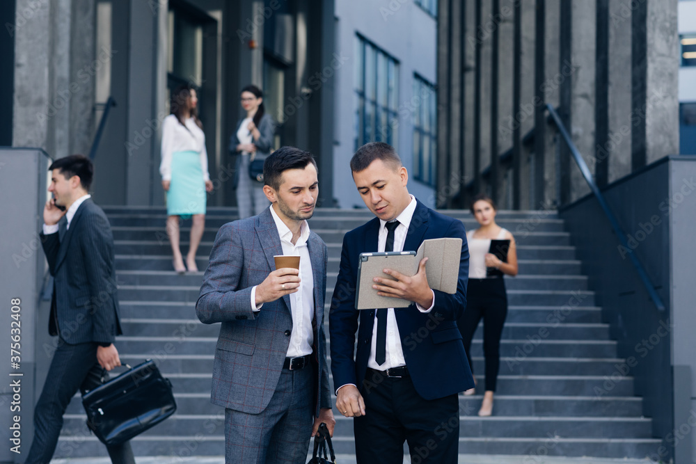 Two Caucasian handsome businessmen in suits and ties holding coffee in hands and walking from office building after work day. Stylish men talking and discussing business Two Caucasian handsome businessmen in suits and ties holding coffee in hands and walking from office building after work day. Stylish men talking and discussing business