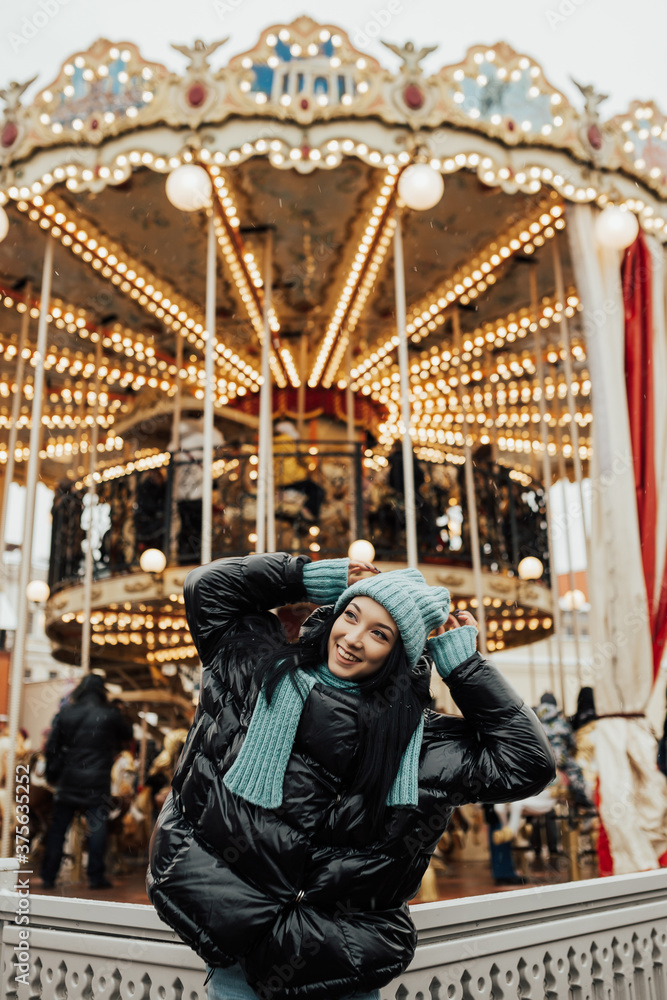 Beautiful smiling woman posing near carousel in Christmas market. Outdoor photo of happy dark-haired girl relaxing in amusement park in winter. Street style. Beautiful smiling woman posing near carousel in Christmas market. Outdoor photo of happy dark-haired girl relaxing in amusement park in winter. Street style.