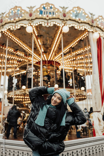 Beautiful smiling woman posing near carousel in Christmas market. Outdoor photo of happy dark-haired girl relaxing in amusement park in winter. Street style.