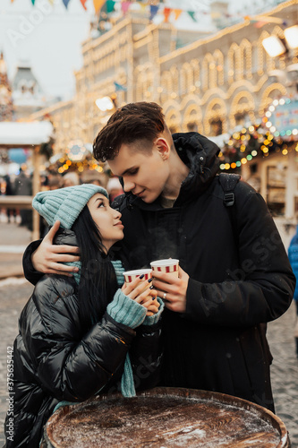 Happy couple in warm clothes drinking coffee on a Christmas market. They looking at each other and smiling. Winter holidays, hot drinks and people concept.