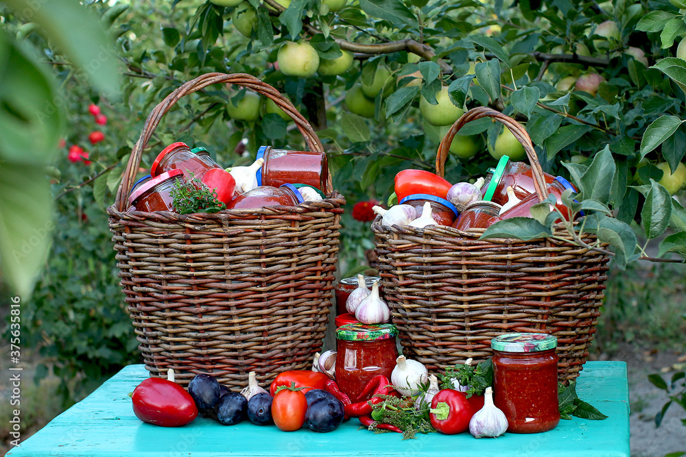 Two wicker baskets full of preservation for the winter on the background of vegetables on a table in the garden. Plum sauce, red bell pepper, garlic, basil, mint, chili. Stocks for the winter. 