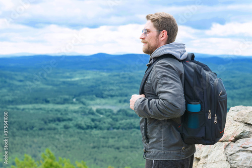 The guy stands on the top of the mountain with a backpack and enjoys the view.