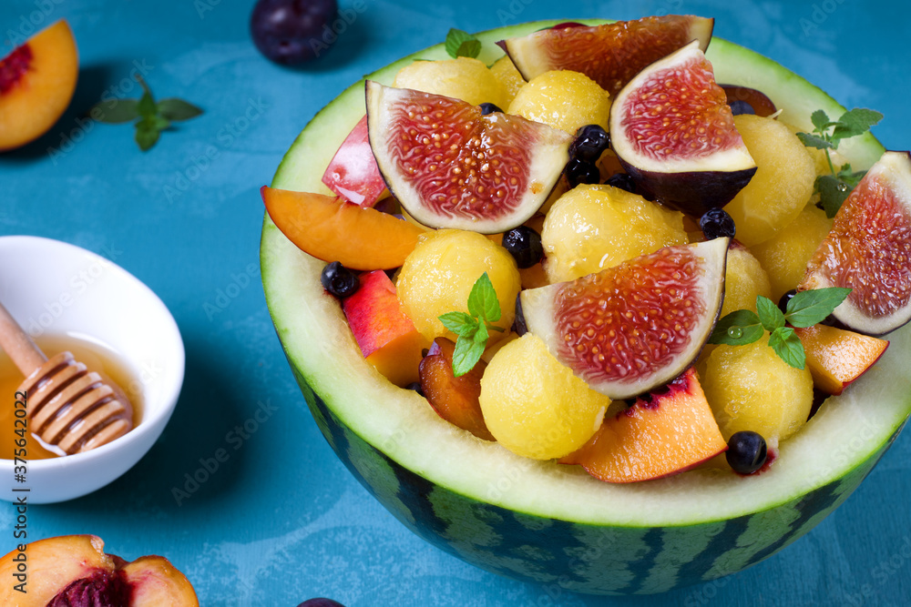 Fruit salad with yellow watermelon, fig, nectarine, plum, bilberry, apple, banana and honey in the watermelon half on the blue table