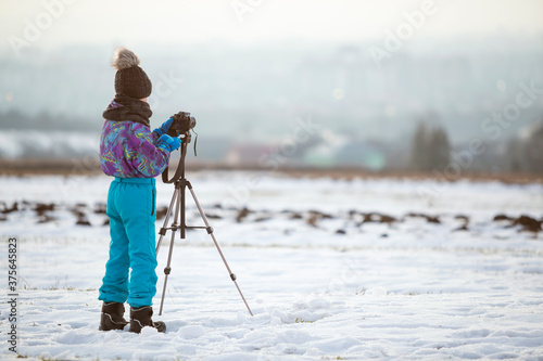 Child boy taking pictures outside in winter with photo camera on a tripod on snow covered field.