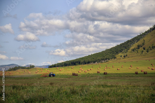 Rural autumn landscape. A tractor on a green field collects hay into bales. In the background there are beautiful mountains  a blue sky with white clouds. Harvesting  agriculture.