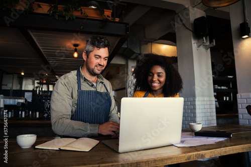 Male and mixed race female coffee shop owners smiling at laptop in coffee shop.