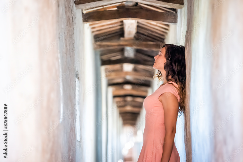 Fototapeta premium Young woman in light pink dress side profile standing leaning on wall of fortress tunnel passage in Castiglione del Lago in Italy