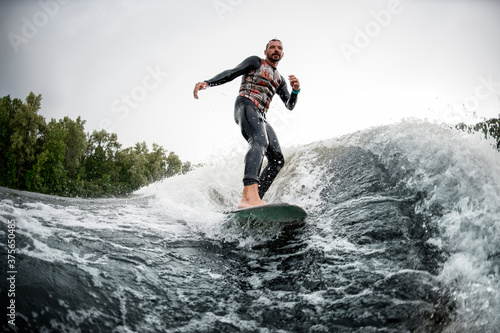 Sporty man rides surfboard on the waves behind boat.