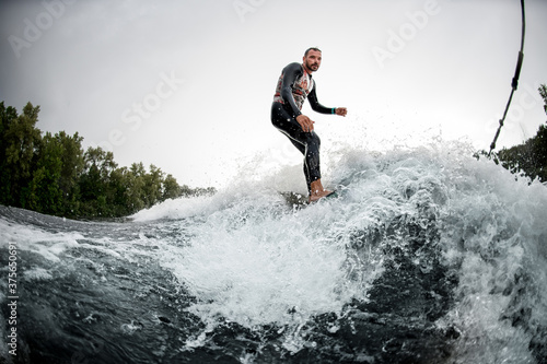 Brutal sporty man rides surfboard on the waves behind boat.