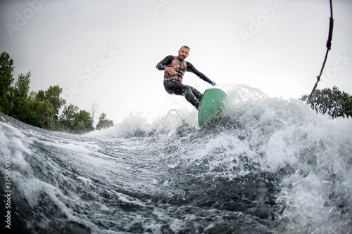 view on cheerful surfer who riding foaming river wave from motorboat at sunny day.