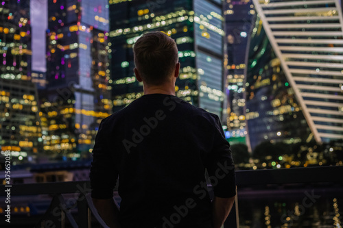 A business man in a black sweater stands with his back to the camera with a view of the night illumination business skyscrapers in Moscow