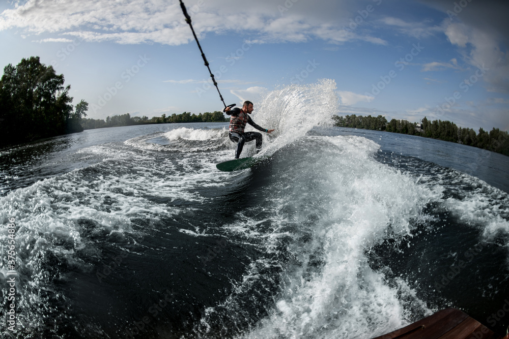 Obraz premium sports man holds stretched rope from motor boat and rides on wakeboard on river water