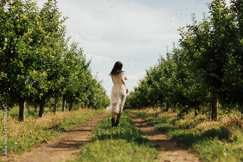 Girl runs through a apple garden in the summer.  Happy young girl in a white dress running in the apple garden. Trees full of ripe fruits. Back view. © eduard