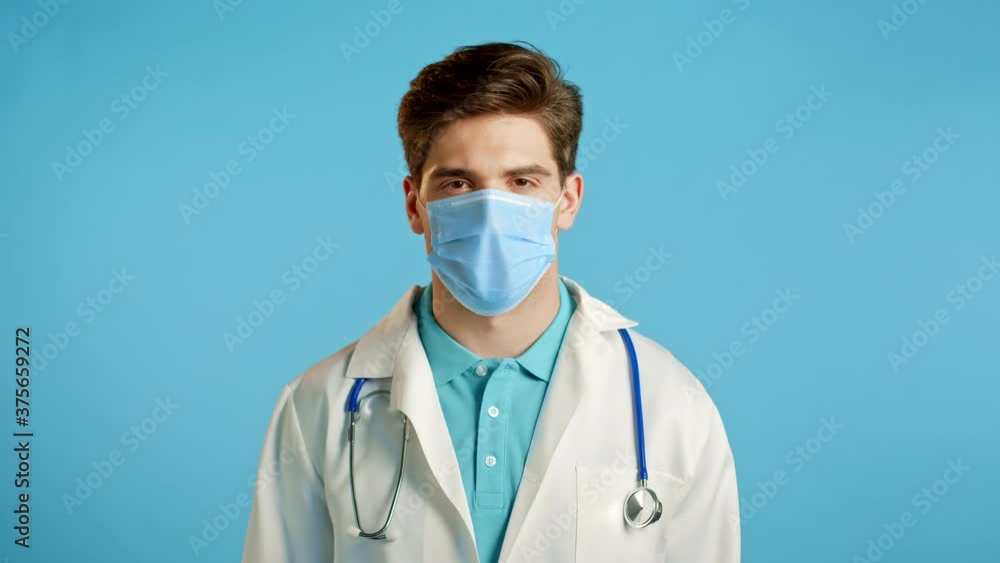 Portrait of satisfied doctor in professional medical mask and coat showing yes sign by head. Man permits, treats approvingly. Doc man isolated on blue background. Portrait of satisfied doctor in professional medical mask and coat showing yes sign by head. Man permits, treats approvingly. Doc man isolated on blue background.