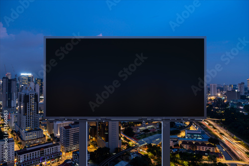 Blank black road billboard with Kuala Lumpur cityscape background at night time. Street advertising poster, mock up, 3D rendering. Front view. Concept of marketing to promote or sell idea or product.