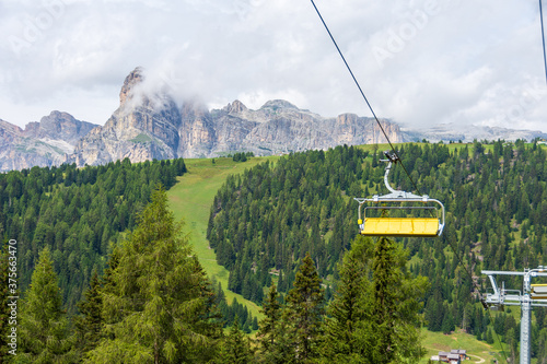 Yellow chairlift connecting valley floor to high peaks of Italian Alps. Sassongher Dolomite mountain is visible in the background, surrounded by green meadows. Colfosco - Val Badia, Italy
