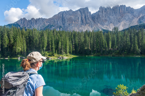 A young woman wearing a face mask admiring a wonderful mountain panorama. In front of her the turquoise water of an Alpine lake and the high peaks of the Latemar mountain group. Carezza Lake, Italy © Travelvolo