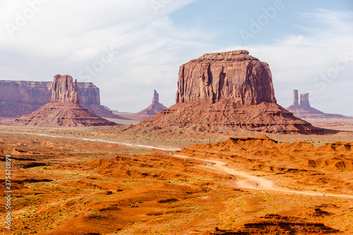 Picture of iconic Merrick Butte rock formation in Oljato-Monument Valley taken on a summer cloudy day from John Ford's point. The scenic drive dirt road with tourist cars winds through the red sand.