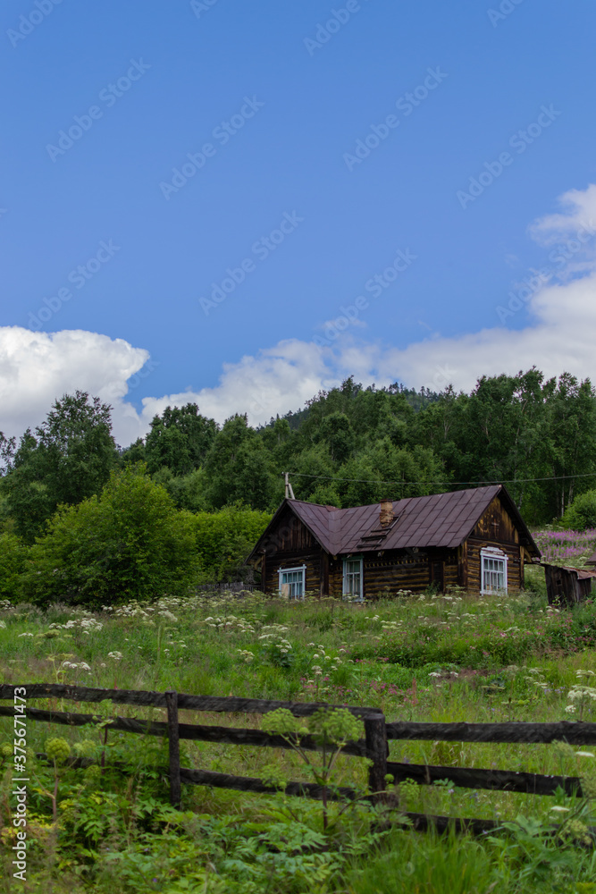 Old wooden brown rustic house stands near a green forest, next to a lawn and a fence, blue sky with clouds backgroung Old wooden brown rustic house stands near a green forest, next to a lawn and a fence, blue sky with clouds backgroung