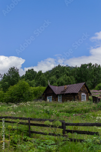 Old wooden brown rustic house stands near a green forest, next to a lawn and a fence, blue sky with clouds backgroung