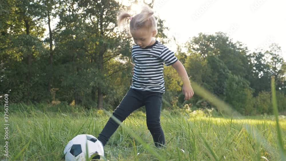 Little girl plays with a soccer ball in the park. The child dreams of football. Happy child kicks a ball on a green lawn. Dream concept, happy childhood. Kid in the park outdoors.