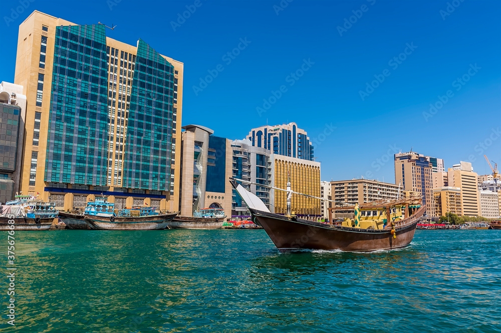 A traditional dhow cargo vessel cruises past modern buildings lining the Dubai Creek in the UAE in springtime