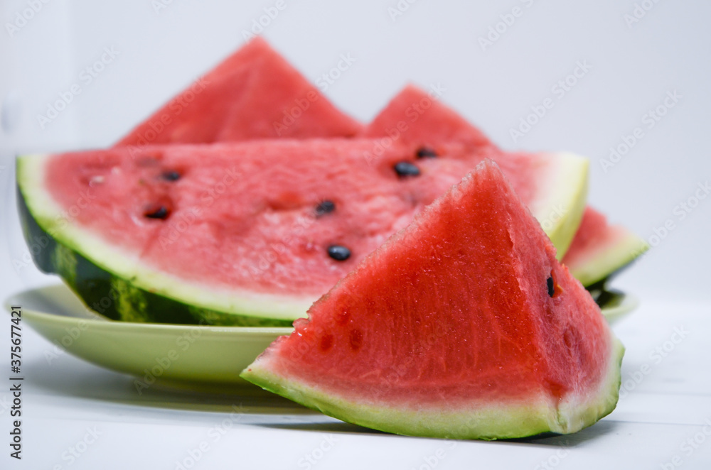 Close-up of triangular piece of watermelon and a plate of watermelon slices on white background. Ripe juicy watermelon, organic farm products, seasonal food.