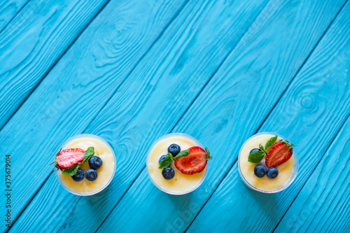 Three light tender desserts with lemon curd, decorated with slices of strawberries, blueberries and mint leaves in plastic transparent glasses on a wooden blue background. Vegan breakfast