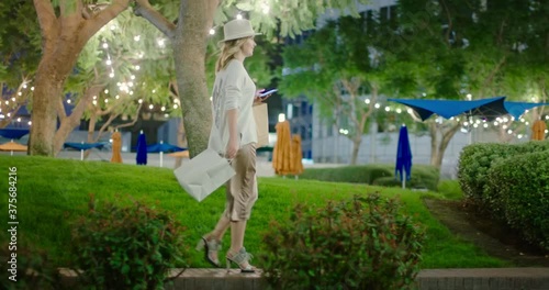 Pretty woman with shopping bags walking by green park on summer evening. Slow motion 4K shopping. Happy woman in stylish dress with shopping bags in downtown with night illumination on background, USA photo