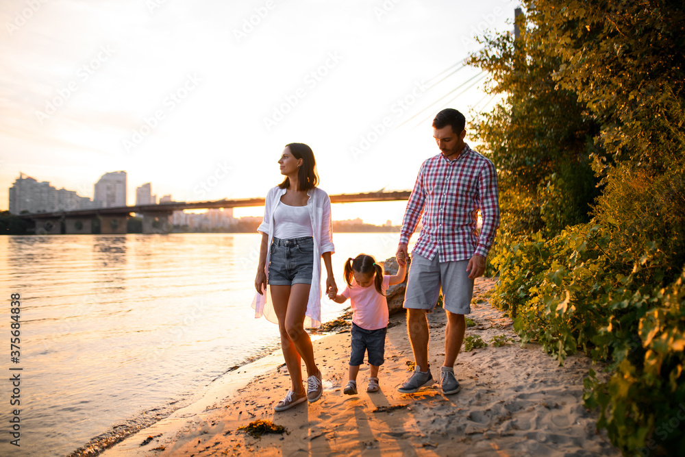 Young family couple and little daughter are walking along the sandy river bank