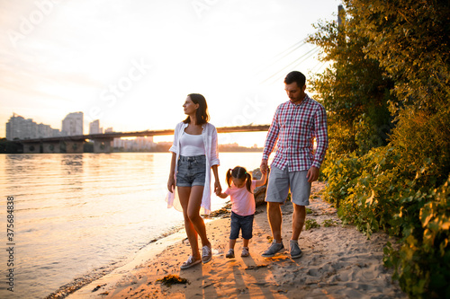 Young family couple and little daughter are walking along the sandy river bank