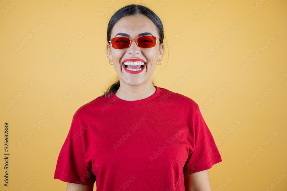 Young beautiful woman wearing red sunglasses over isolated yellow background smiling and looking at the camera. Young beautiful woman wearing red sunglasses over isolated yellow background smiling and looking at the camera.