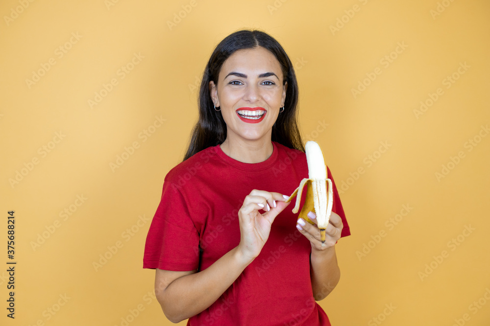 Young beautiful woman wearing casual red t-shirt on yellow background holding a banana.
