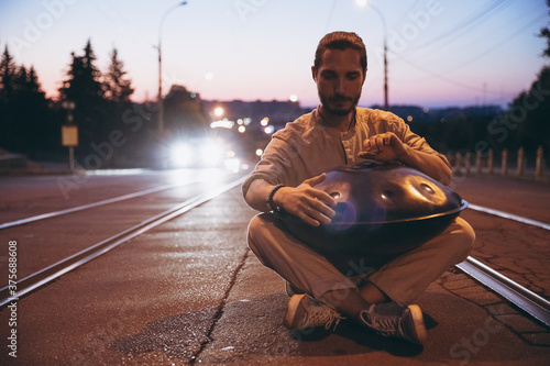 young bearded man playing handpan while sitting on the road, musical harmony in the middle of an urban city. Copy space photo