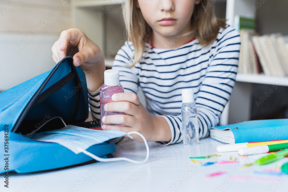 Little girl packing blue backpack in kids's room. Face mask, bottle of sanitizer, stationery, pens, multicolored markers, notebooks. Back to school. Mom's hygiene,safety precautions after coronavirus Little girl packing blue backpack in kids's room. Face mask, bottle of sanitizer, stationery, pens, multicolored markers, notebooks. Back to school. Mom's hygiene,safety precautions after coronavirus