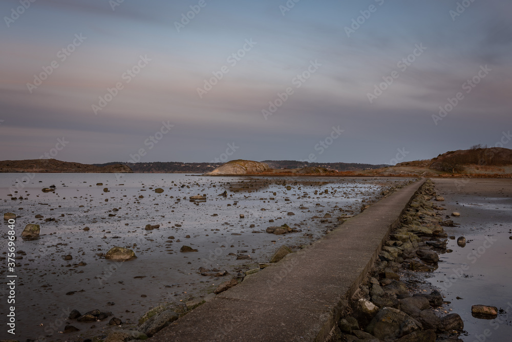 A concrete pathway that leads to a small island in low tide. Picture is shot in early morning light. Stones are visible on the sea bed because of the low water level. It is a calm and relaxing photo.