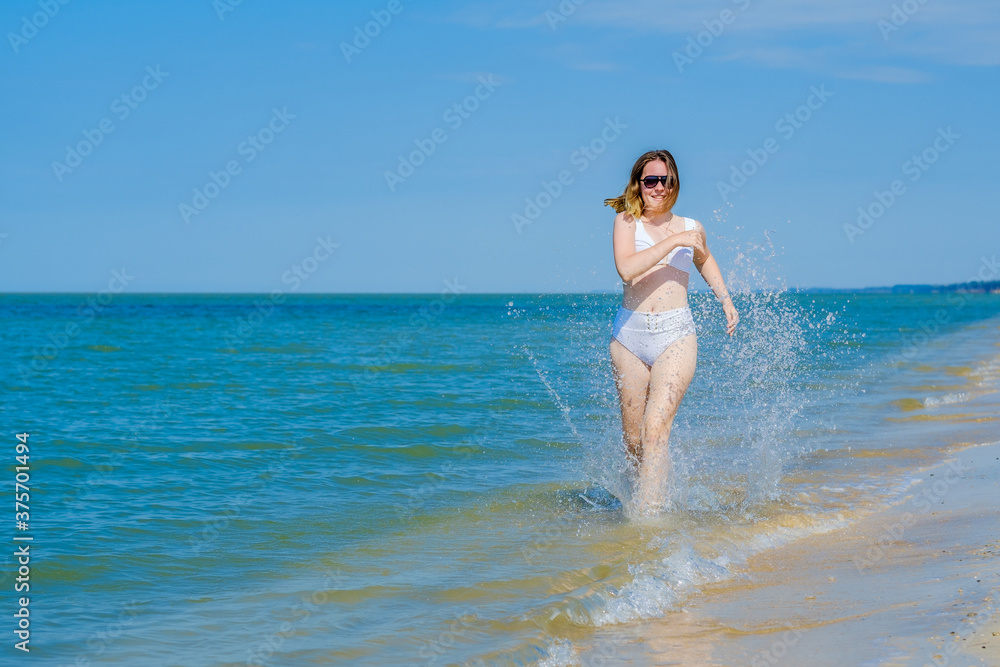 A young girl runs along the sandy sea beach along the surf. Splashes of sea water. The girl is wearing a white swimsuit and sunglasses. Freedom and carelessness. Outdoor fitness. Sunny day. Copy space A young girl runs along the sandy sea beach along the surf. Splashes of sea water. The girl is wearing a white swimsuit and sunglasses. Freedom and carelessness. Outdoor fitness. Sunny day. Copy space