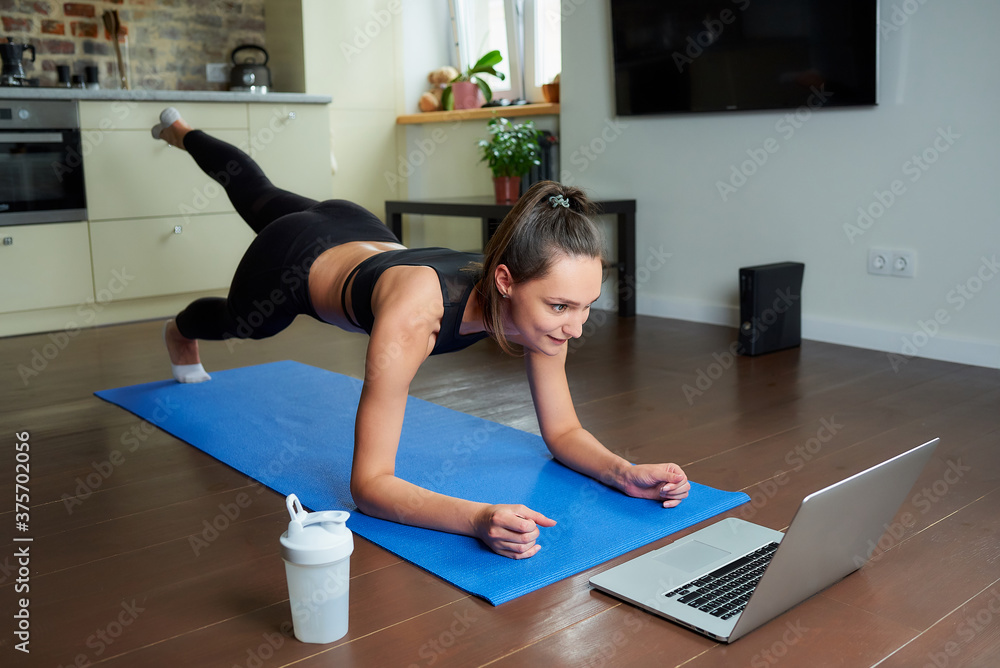 A sporty girl in a workout tight suit is doing buttocks workout with an online video on a laptop. A female coach conducting a remote fitness class on the blue yoga mat at home. A sporty girl in a workout tight suit is doing buttocks workout with an online video on a laptop. A female coach conducting a remote fitness class on the blue yoga mat at home.