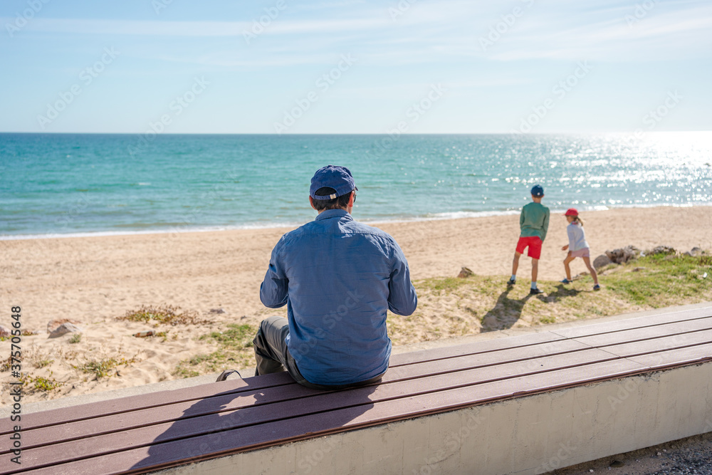 Father with children relaxing on a bench near sea beach, sunshine outdoors background. Back view of people dreaming romantic image.