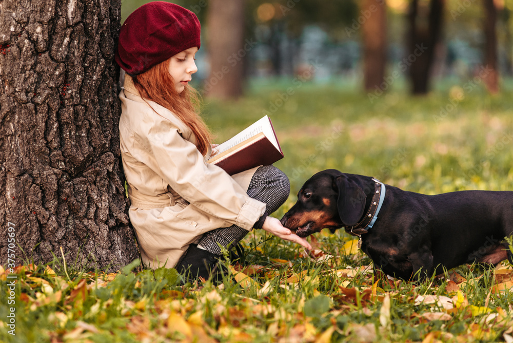 A cute little girl is reading a book sitting under a tree in the park ...