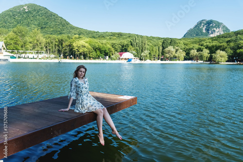 A beautiful and sexy woman, with long hair in a summer dress, spends time on the shore of the lake, surrounded by forests and mountains. Rest and relaxation concept