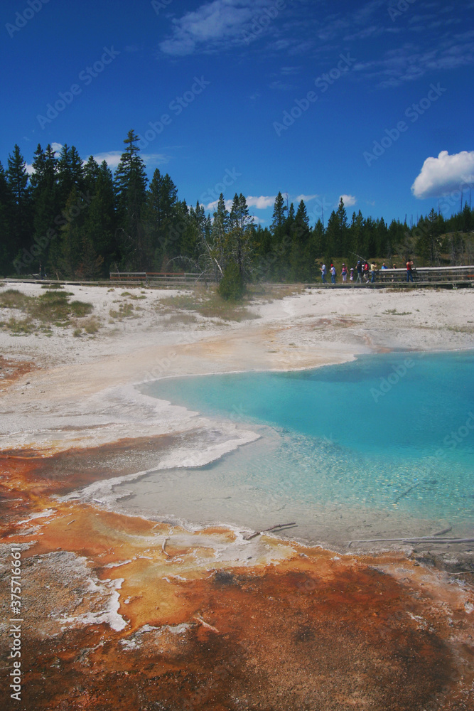 Distant tourists view a brilliant blue colored geothermal hot spring with a rust colored edge, very typical colors to see in Yellowstone's landscapes. Distant tourists view a brilliant blue colored geothermal hot spring with a rust colored edge, very typical colors to see in Yellowstone's landscapes.