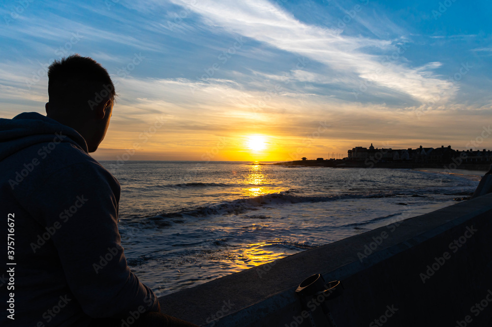Young male sat on wall over looking sunset at porthcawl wales uk, focus on male, blured background