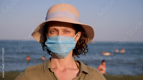 Portrait of beautiful woman in panama wearing disposable mask. Tender lady in surgical mask standing on crowdy beach. Seascape with swimmers on background. Protection on vacation during coronavirus photo