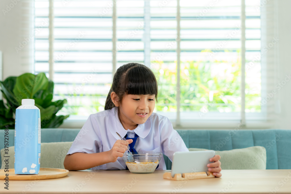 Asian elementary school student girl eating breakfast cereals with milk and looking cartoon in smartphone to distractions to eat it in morning school routine for day in life getting ready for school. Asian elementary school student girl eating breakfast cereals with milk and looking cartoon in smartphone to distractions to eat it in morning school routine for day in life getting ready for school.