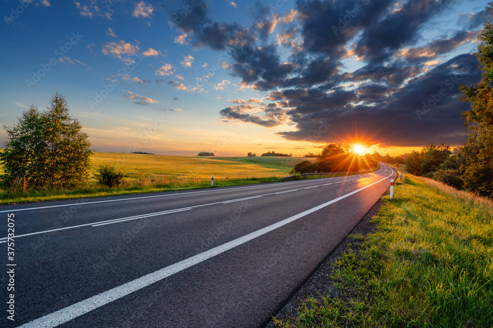 Empty asphalt road in rural landscape in the rays of the sunset with dark storm cloud