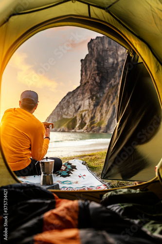 Camping with tent in Lofoten islands, Northern Norway, Kvalvika beach, during sunset. Person sitting in front of a tent holding a hot drink mug.