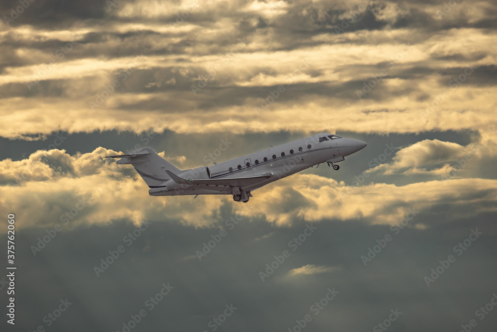 Fototapeta premium White small private turbofan-powered twin-engine business jet airplane flying in air after takeoff from airport. Dramatic orange grey cloudy sky at sunset after rain storm, dynamic weather conditions.